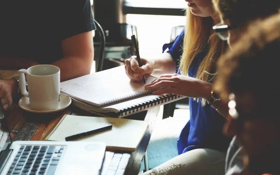 Pessoas em reunião, com cadernos, notebook e café na mesa
